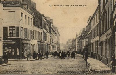 On peut voir à ce carrefour avec la rue du Lycée, une épicerie-tabac, ce qui était trés fréquent à l'époque. Les pavés qui recouvraient la chaussée de nombreuses rues de Saint-Omer sont bien visibles sur cette carte. Si vous vous baladez aujourd'hui dans ce lieu, vous verrez qu'ils sont encore présents au sol.